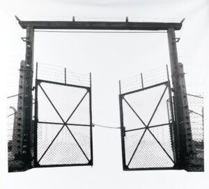 Monumental film negative photograph titled "Barbed Gate" by artist Casey McKee, depicting an inverted light-and-shadow view of the historic gate at the Auschwitz II-Birkenau concentration camp.